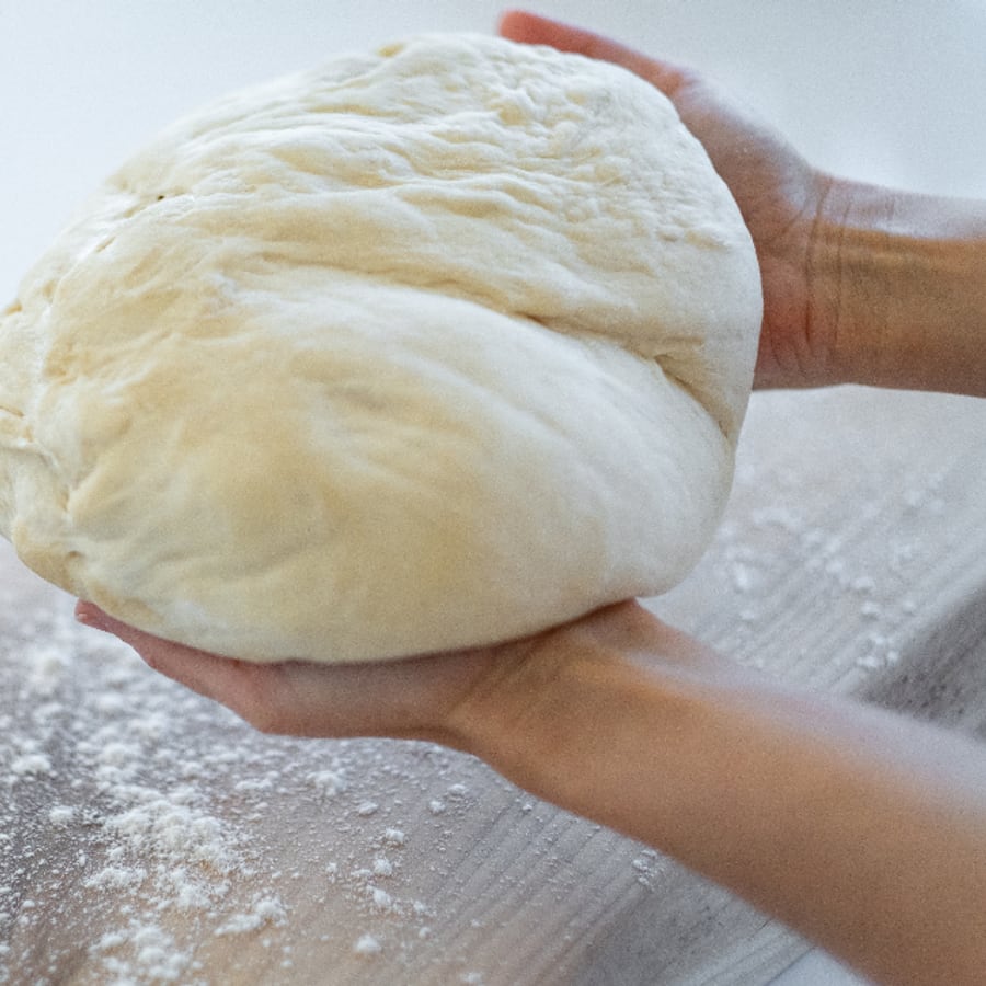 Twee handen houden een bol pizzadeeg boven een met bloem bestoven werkblad